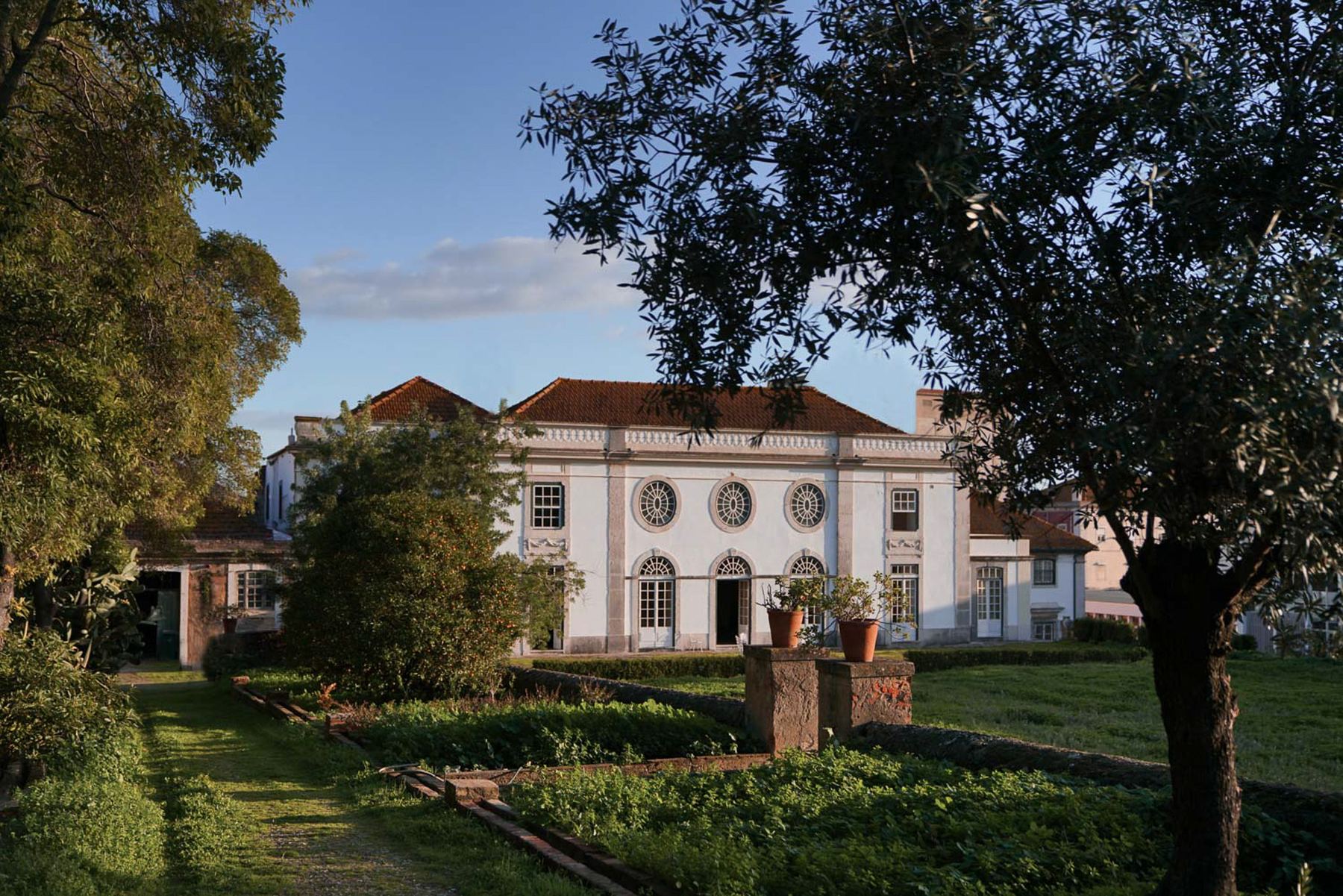 Vista exterior do Palácio do Grilo em Lisboa, com a sua fachada barroca branca e jardins verdejantes ao pôr do sol.