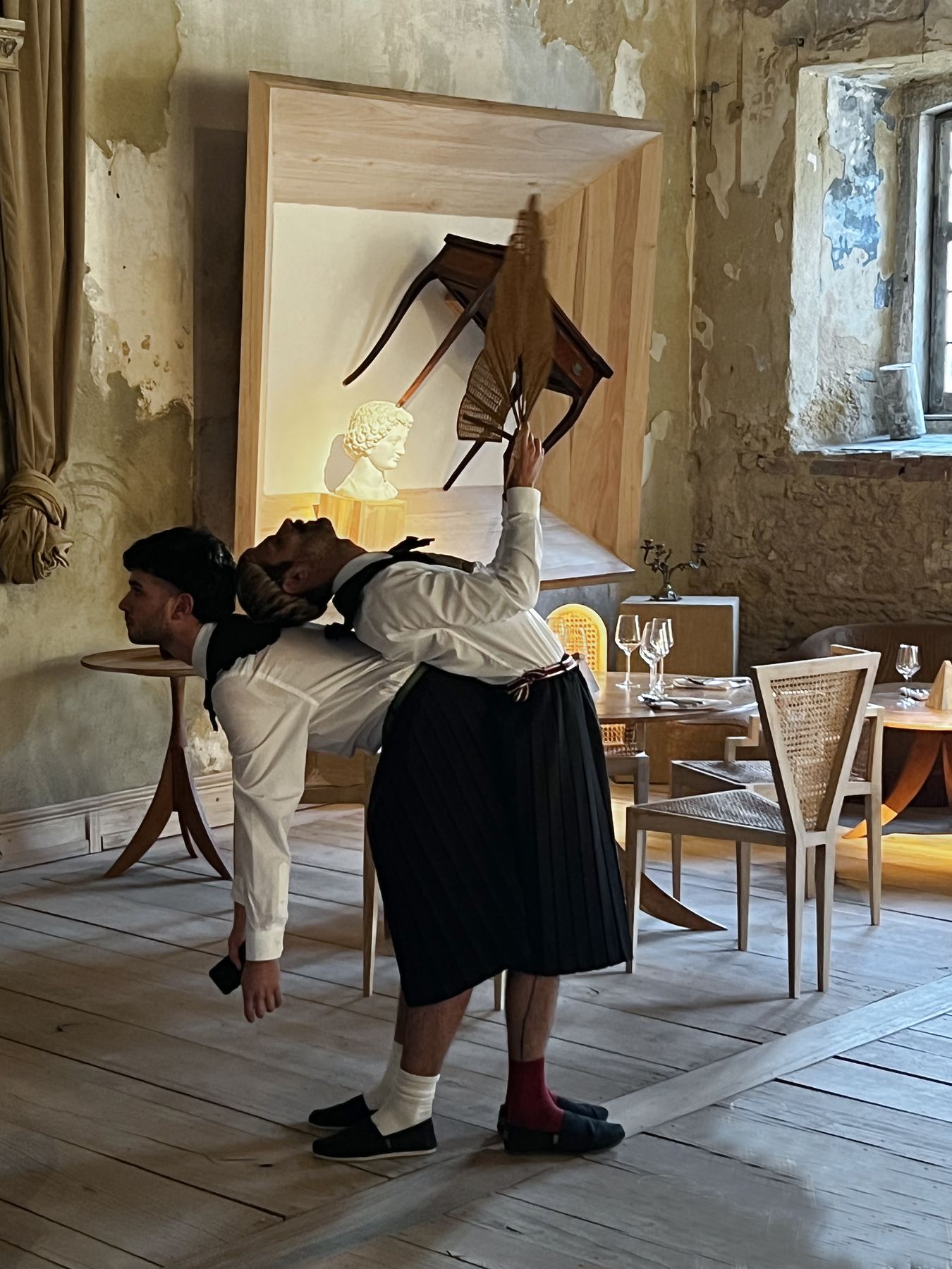 Duas figuras performáticas interagem no interior rústico-chique do Palácio do Grilo em Lisboa, com uma instalação de arte de fundo.