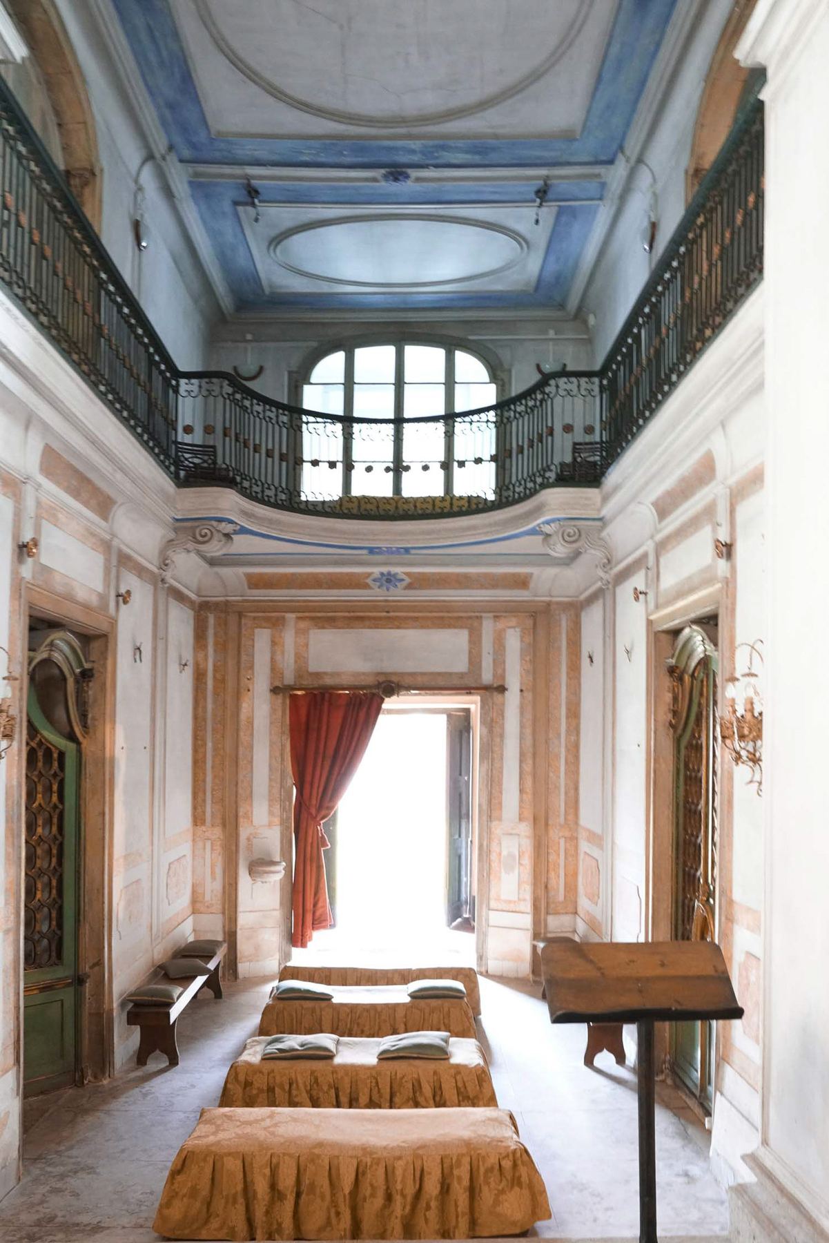 Um grande salão com paredes pintadas e teto barroco, com uma varanda de ferro forjado e quatro camas baixas alinhadas no Palácio do Grilo, Lisboa.