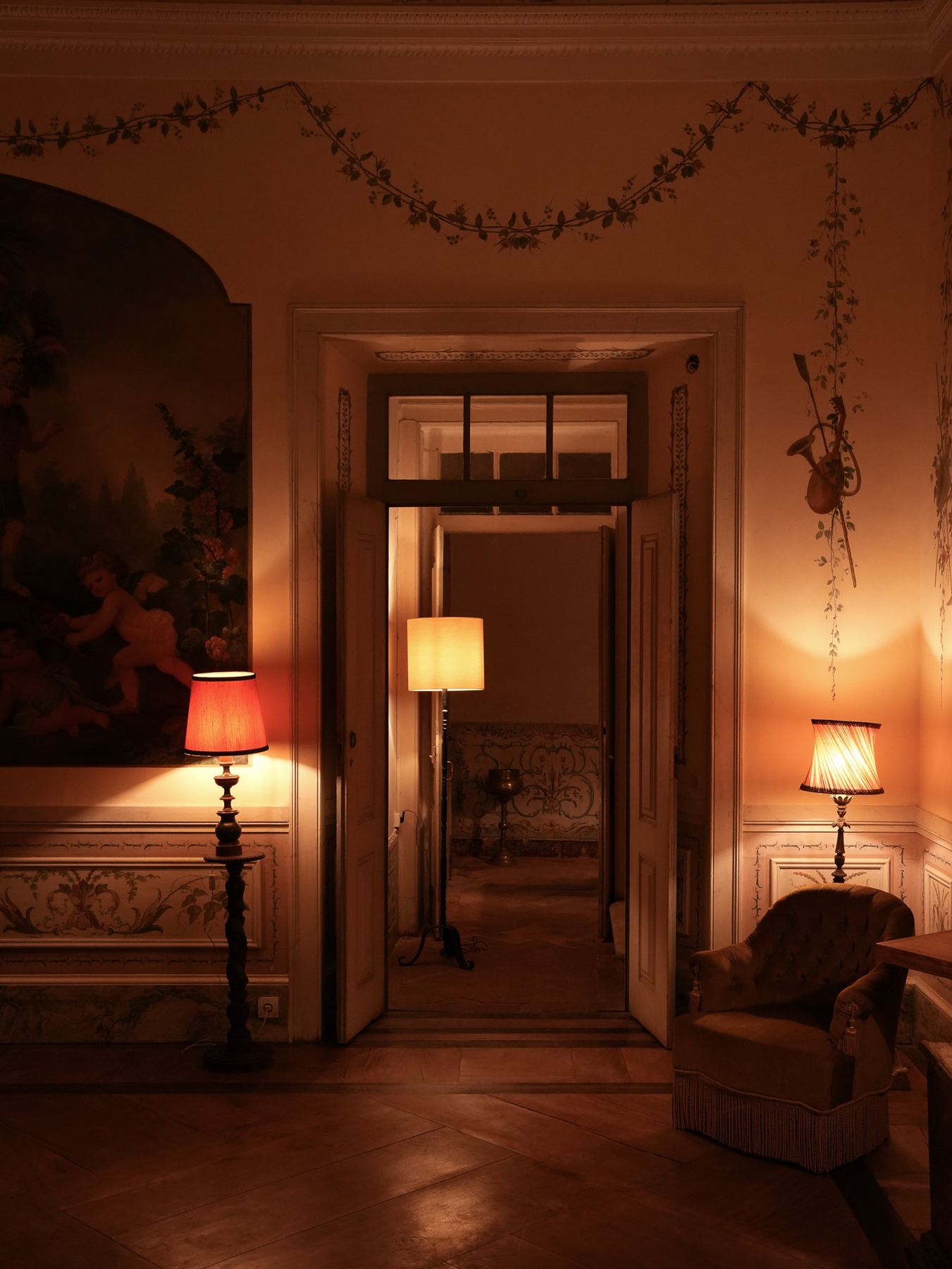 Um corredor atmosférico no Palácio do Grilo, em Lisboa, revela chão de madeira, arte barroca e candeeiros acesos que criam um ambiente acolhedor.