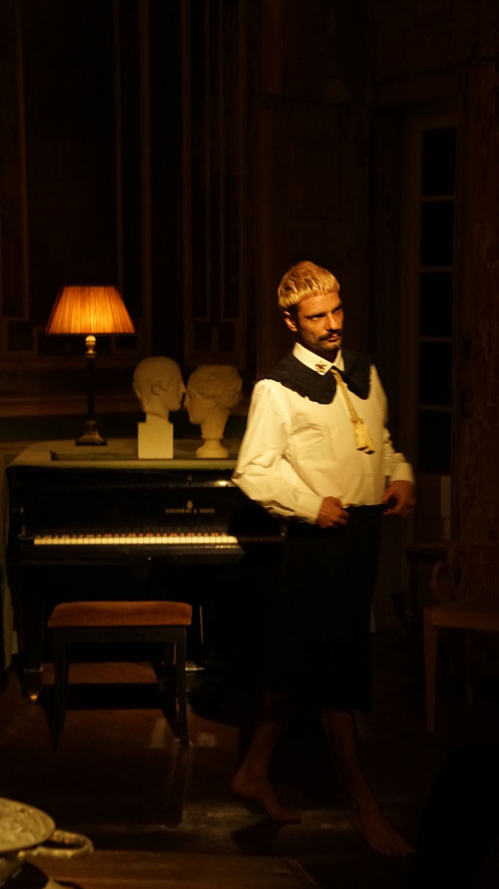 Um performer de cabelo loiro e bigode posa descalço junto a um piano e lâmpada em Palácio do Grilo, Lisboa.