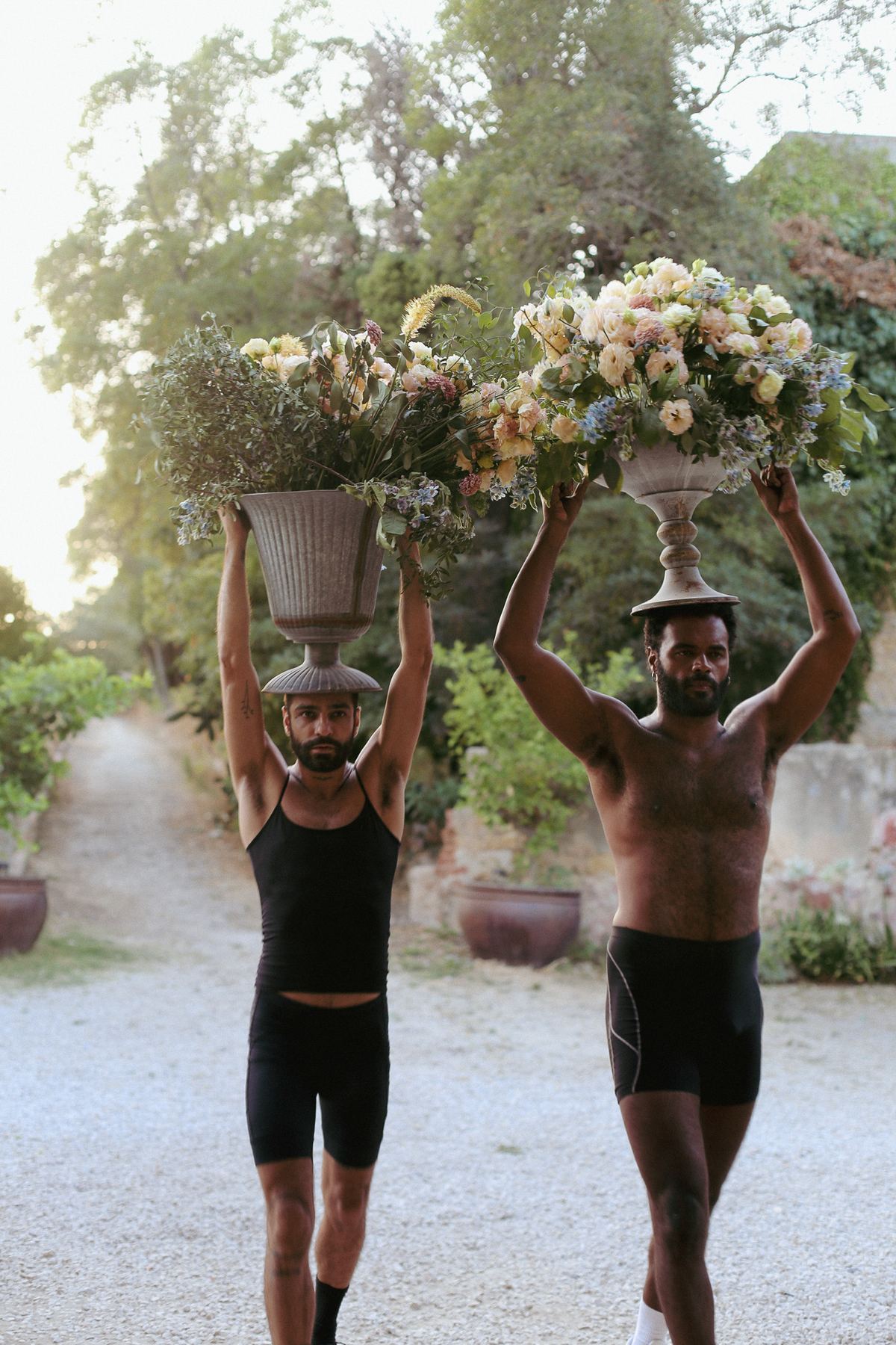 Dois homens carregam vasos floridos na cabeça, uma cena performática nos jardins do Palácio do Grilo, Lisboa.