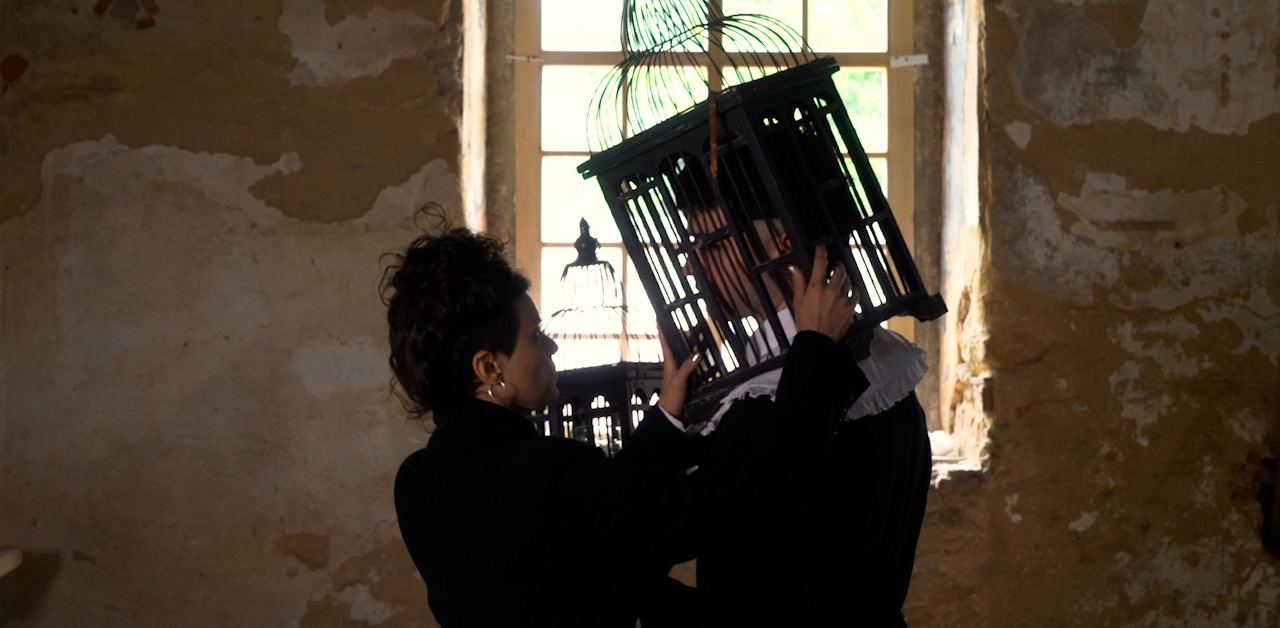 Duas figuras performáticas interagem com uma gaiola de pássaros, iluminadas por uma janela no Palácio do Grilo, Lisboa.