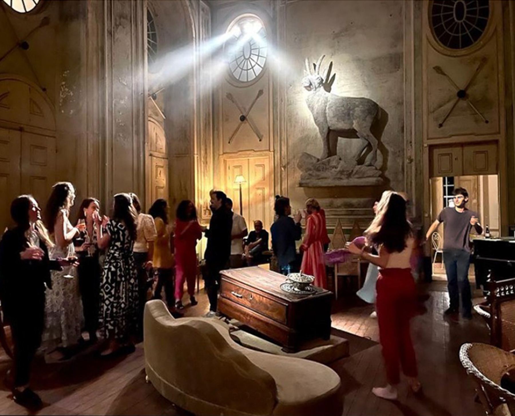Um animado grupo de pessoas socializa num salão barroco do Palácio do Grilo, com uma estátua de veado e mobiliário antigo, em Lisboa.