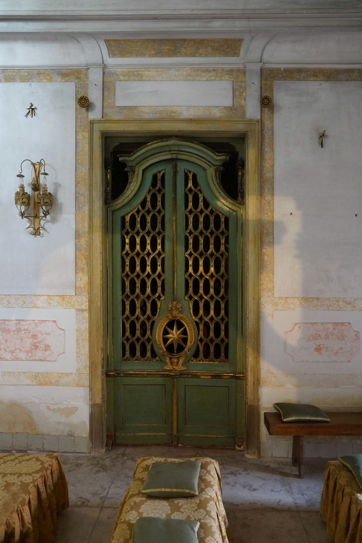 Um detalhe do Palácio do Grilo, com uma porta barroca verde e dourada, paredes claras e bancos elegantemente cobertos.