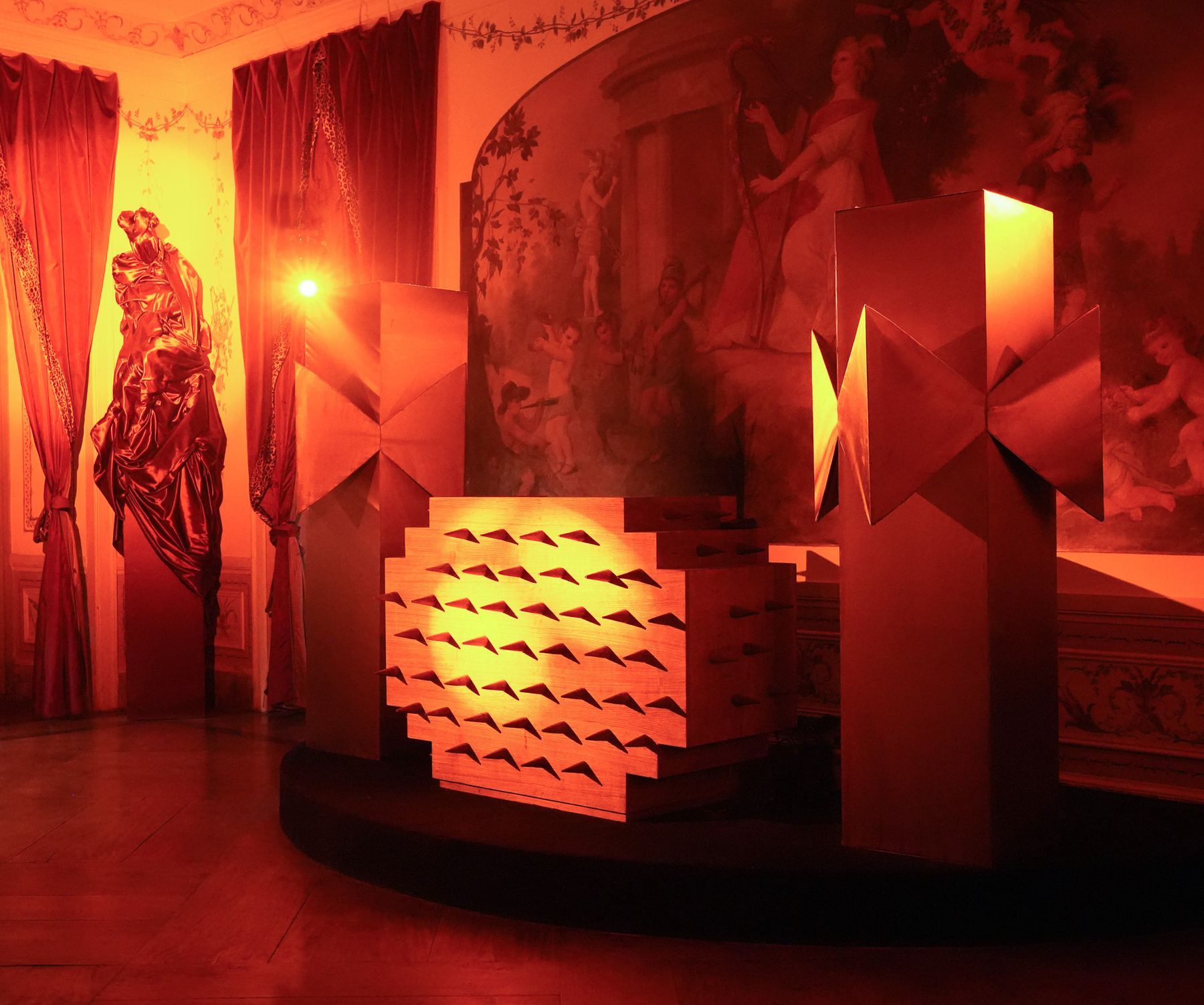 Uma sala do Palácio do Grilo em Lisboa com iluminação vermelha, um mural barroco e instalações geométricas modernas.