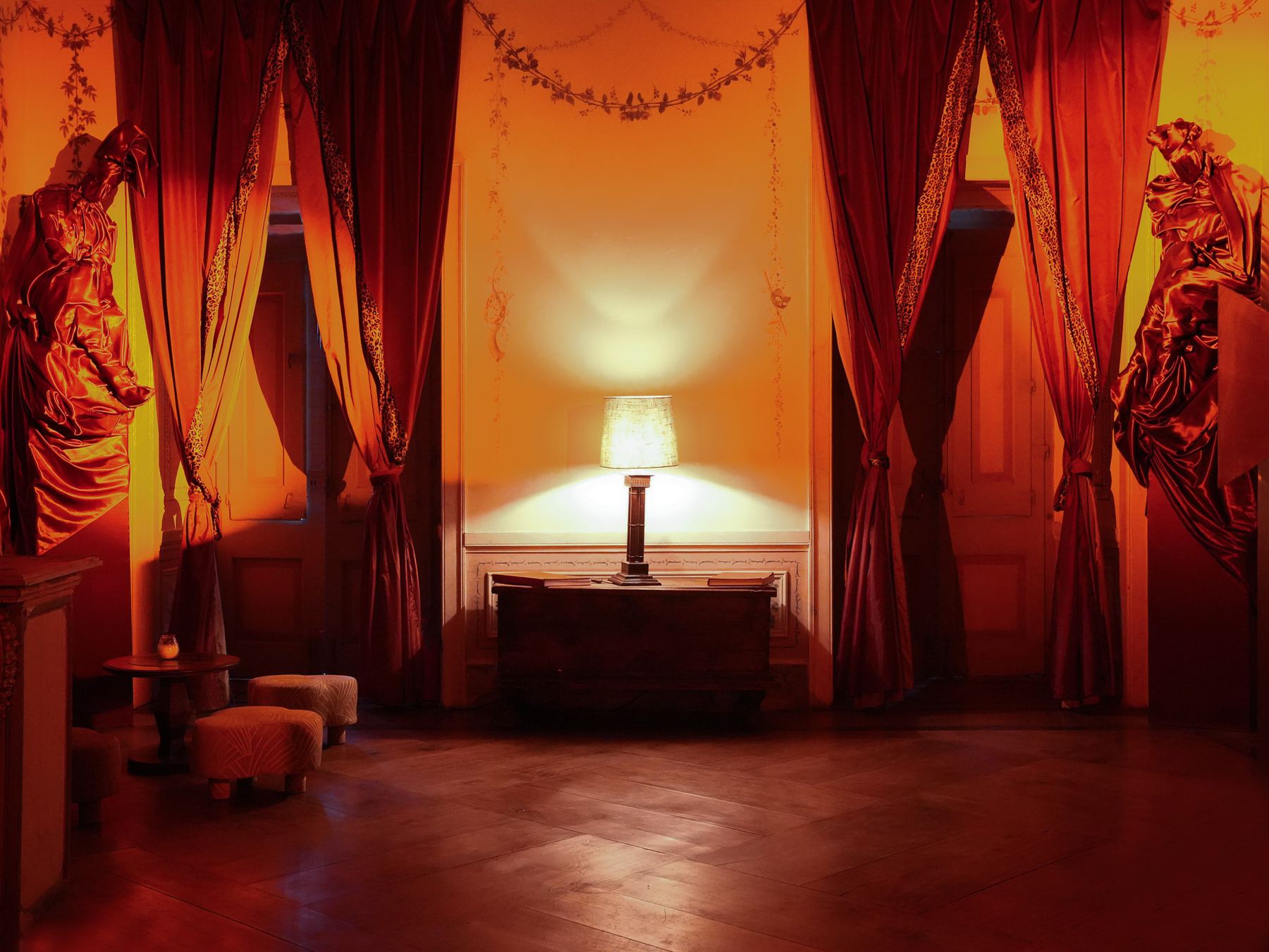 Um quarto de Palácio do Grilo em Lisboa com iluminação âmbar, cortinas de veludo carmesim e duas estátuas drapeadas.