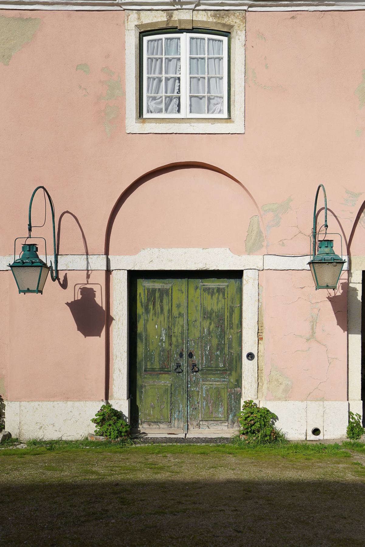 Uma antiga porta verde e desgastada com lanternas e uma janela em Palácio do Grilo, Lisboa, sob luz solar.